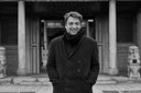 Giorgio Spina in a dark double-breasted coat outside a temple gateway, black-and-white portrait