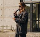 Sara Jowett in sunglasses and an oversized grey blazer speaking into a microphone outdoors
