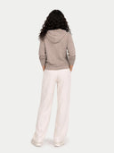 back view of a woman wearing a beige hoodie and white pants on a white background.