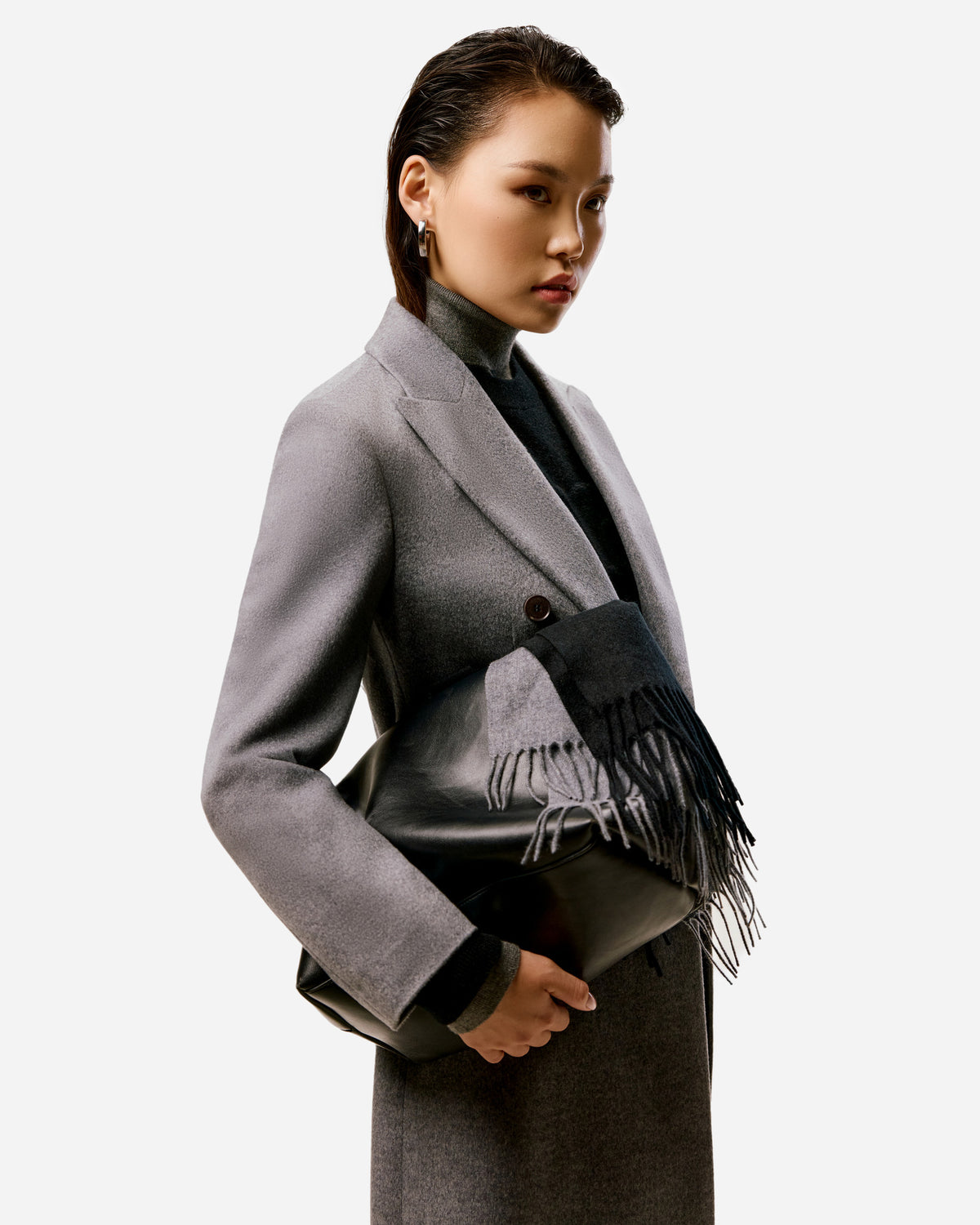 Woman in a grey cashmere coat holding a black leather bag draped with a grey and black fringed scarf