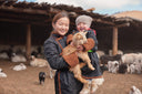 A herder mother in a deel holding her child and a newborn goat kid inside a pen