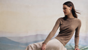 GOBI Cashmere Nomadic Summer campaign — woman in a beige pointelle-knit cashmere sweater and cream pleated skirt seated against a painted steppe landscape