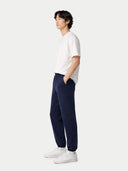 A man in profile wearing a white short-sleeve t-shirt, navy blue jogger pants, and white sneakers, standing against a light gray studio background. #color_ink-blue #ai-image