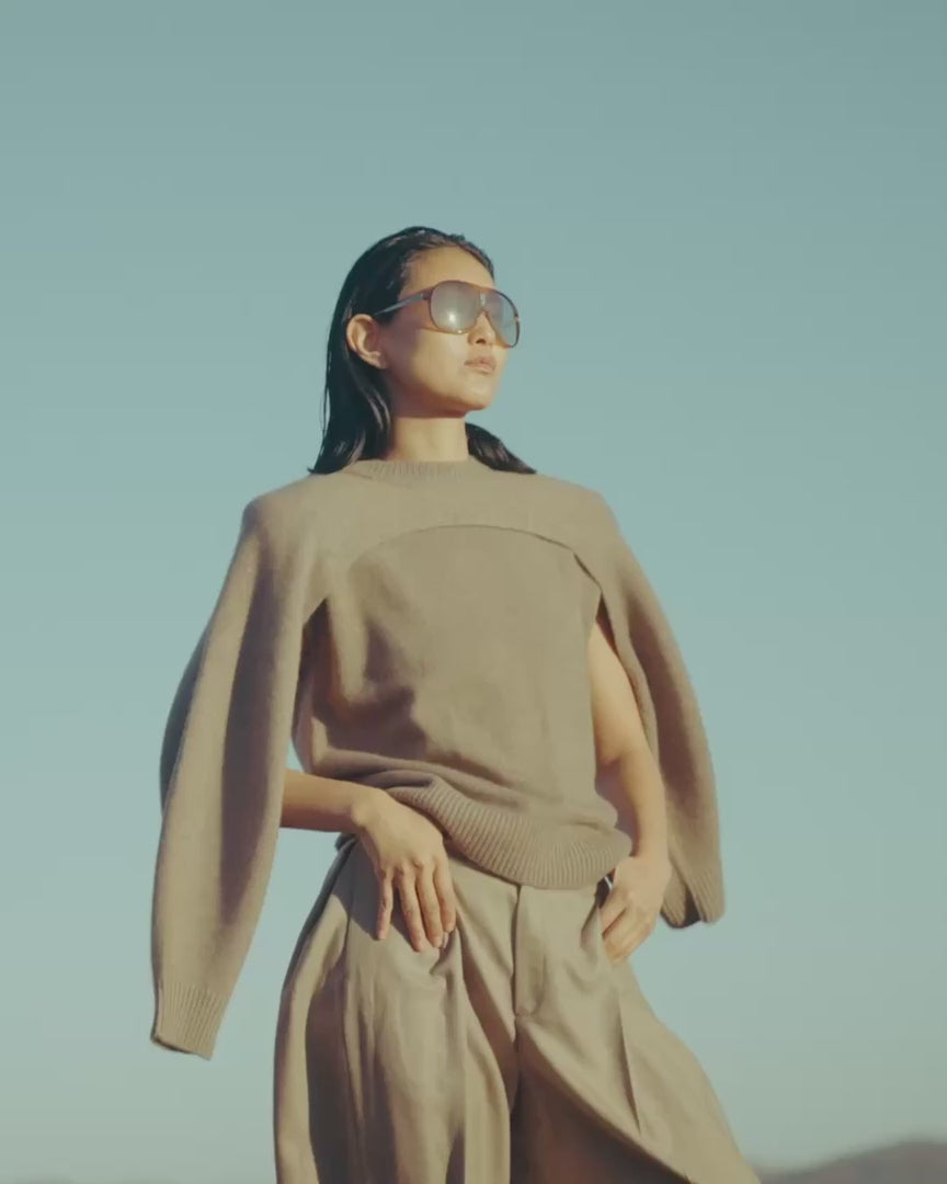 Woman in sunglasses wearing a taupe cashmere knit with a draped shrug and wide-leg trousers against a clear blue sky