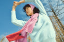 A woman in a mint seamless cashmere sweater and pink scarf stretching her arms against a blue sky