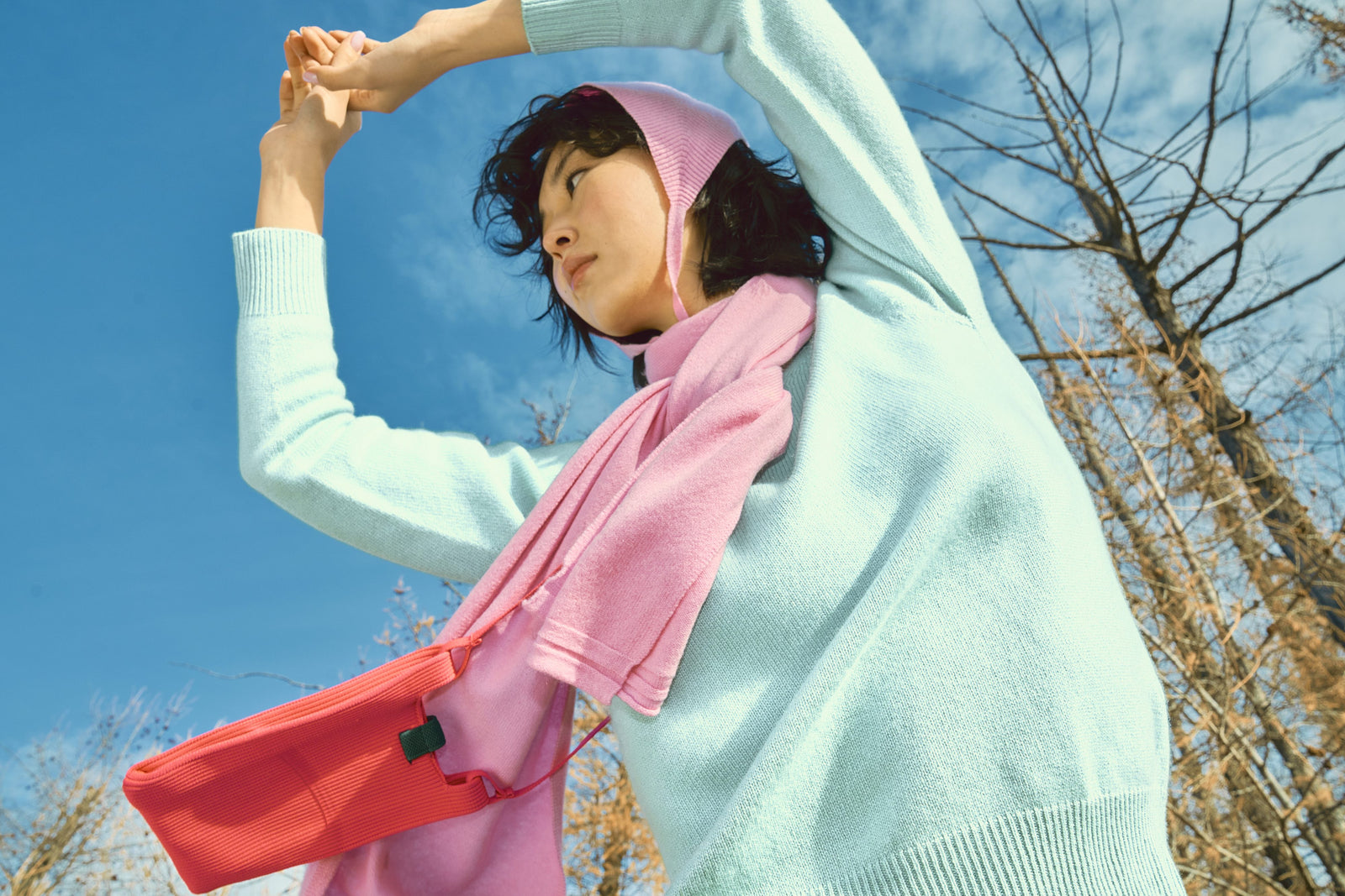 A woman in a mint seamless cashmere sweater and pink scarf stretching her arms against a blue sky
