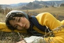 A woman in a yellow cashmere sweater and headscarf lying in autumn grass, hills behind