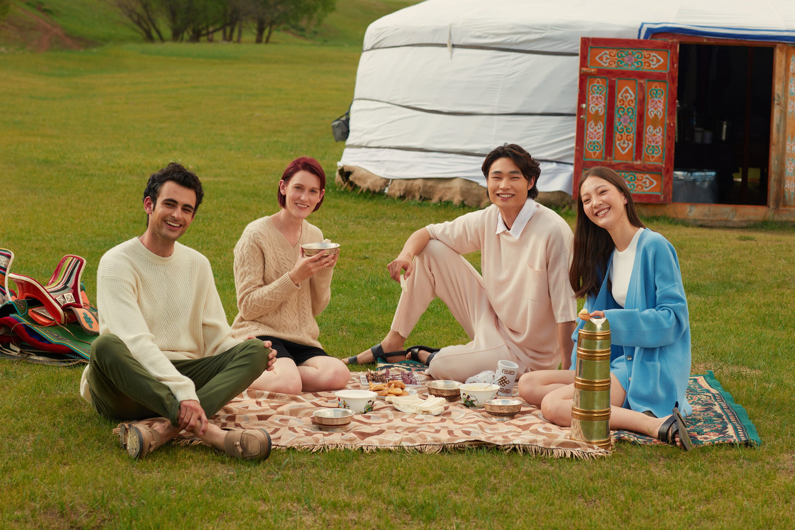 Four friends in cashmere knitwear picnicking on the grass beside a ger