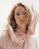 A woman with wind-blown hair in a pale pink cashmere top, a pink cashmere scarf knotted at the neck