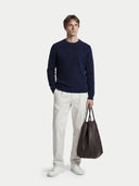 A man in a navy ribbed cashmere sweater and cream trousers carrying a brown leather tote